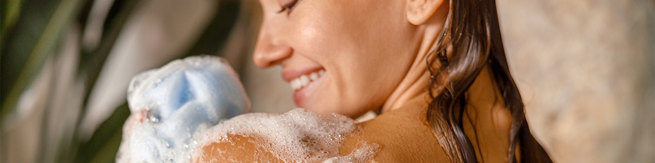 Mulher tomando banho, com espuma de sabonete visível na pele, ilustrando como os sabonetes agem na pele para limpeza e hidratação