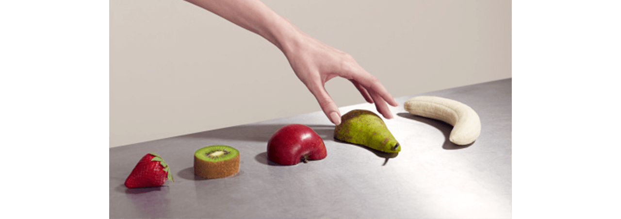 Mão alcançando pera sobre mesa com morango, kiwi, maçã e banana descascada, evocando frescor e o cheiro de frutas maduras.