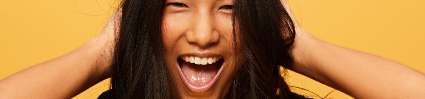 Mulher sorridente sob fundo amarelo vibrante com as mãos nos cabelos saudáveis, ilustrando dicas para evitar cabelo opaco.  