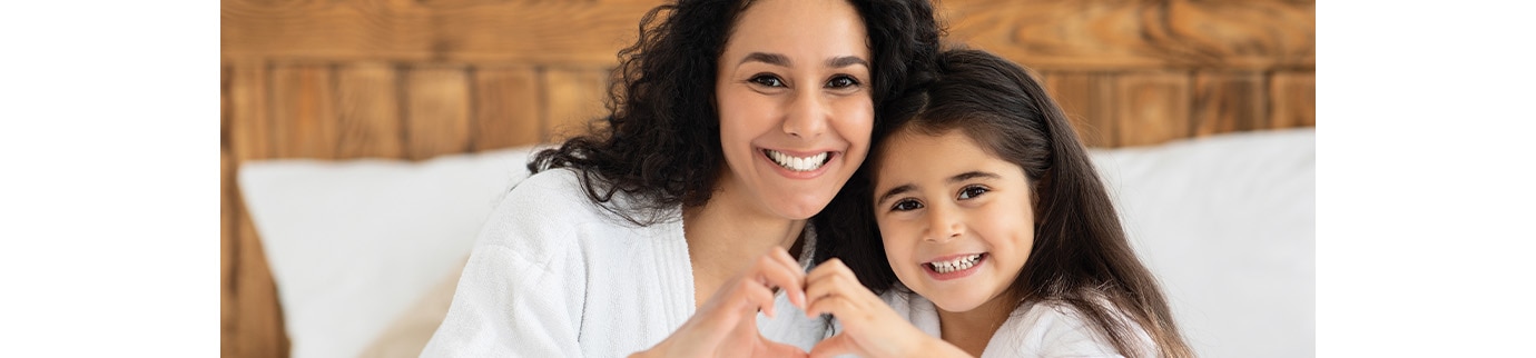 Mãe e filha sentadas na cama usando roupão e formando coração com as mãos, após banho com shampoo e condicionador infantil