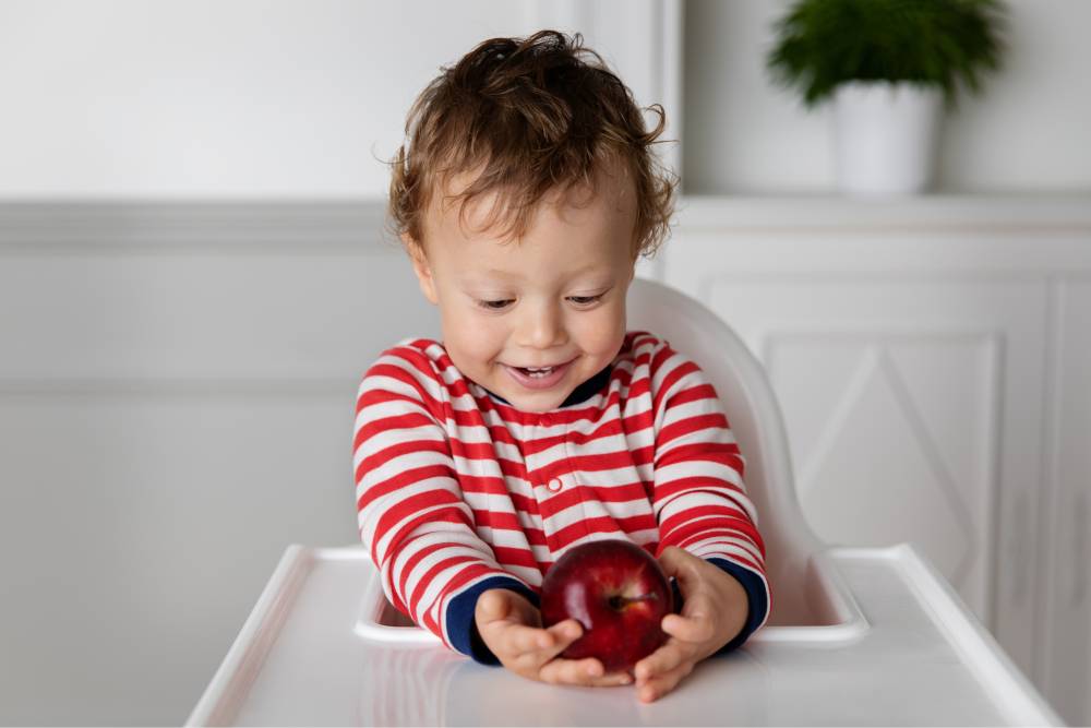 Criança sentada no cadeirão segurando uma maçã e comendo ingredientes naturais