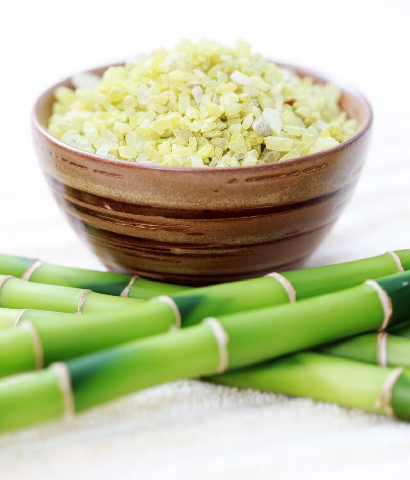 Uma tigela com sais de banho verde com pedaços de bambu a frente, representando o aroma dos sais